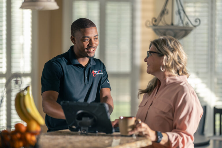 A man in a black polo shirt speaks with a woman in glasses holding a cup at a kitchen counter. A tablet and a fruit bowl are on the counter.