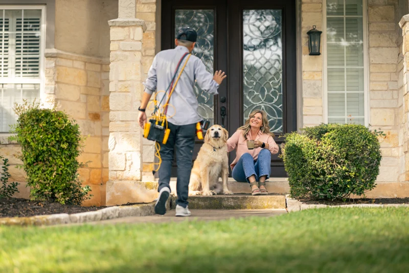 A person with tools approaches a seated woman and a dog on a porch with stone pillars in front of a glass-paneled door.