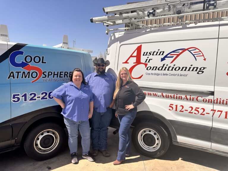 Three people standing between two HVAC service vans, wearing casual work attire and smiling. The vans display logos and contact information for Cool Atmosphere and Austin Air Conditioning.